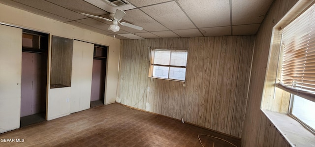 unfurnished bedroom with visible vents, two closets, a drop ceiling, wooden walls, and ceiling fan