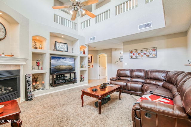 carpeted living room with built in features, a tiled fireplace, ceiling fan, and a high ceiling