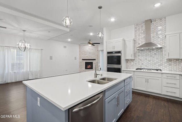 kitchen featuring appliances with stainless steel finishes, ceiling fan with notable chandelier, wall chimney exhaust hood, a fireplace, and sink