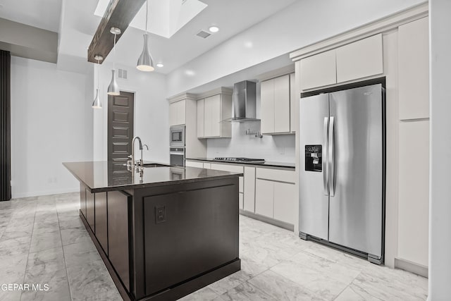 kitchen featuring a skylight, wall chimney exhaust hood, stainless steel appliances, a center island with sink, and pendant lighting