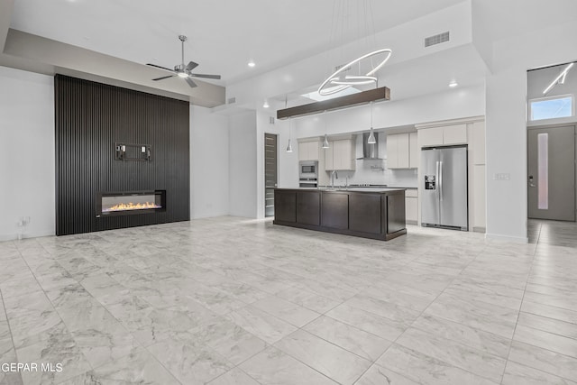 kitchen featuring appliances with stainless steel finishes, ceiling fan, wall chimney range hood, hanging light fixtures, and a kitchen island with sink