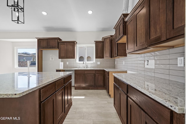 kitchen with light stone countertops, a sink, a center island, dark brown cabinetry, and backsplash