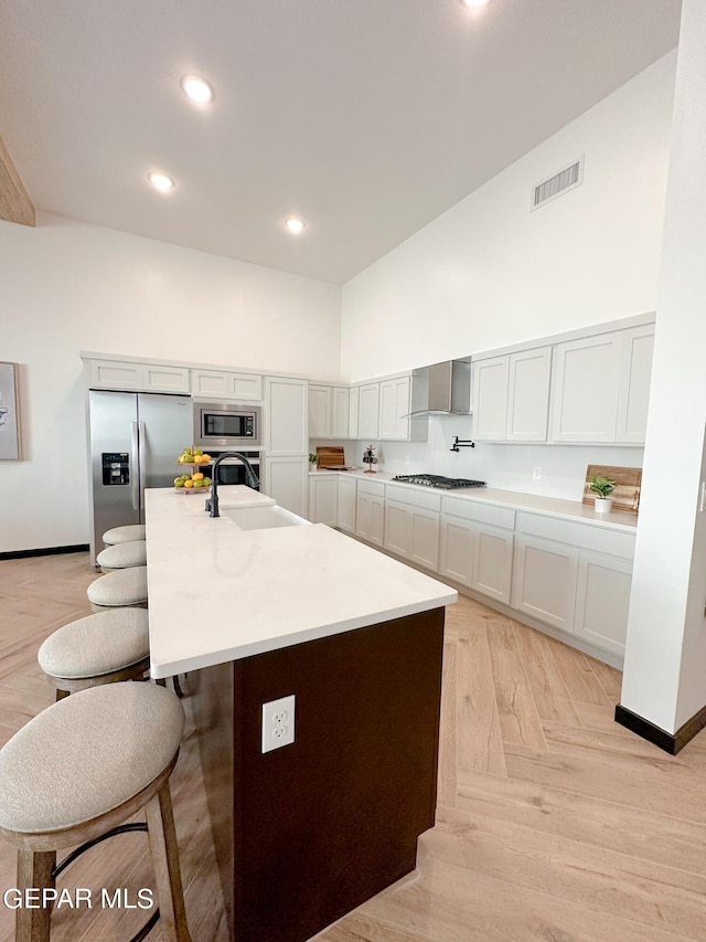 kitchen featuring appliances with stainless steel finishes, wall chimney range hood, white cabinets, an island with sink, and light parquet floors