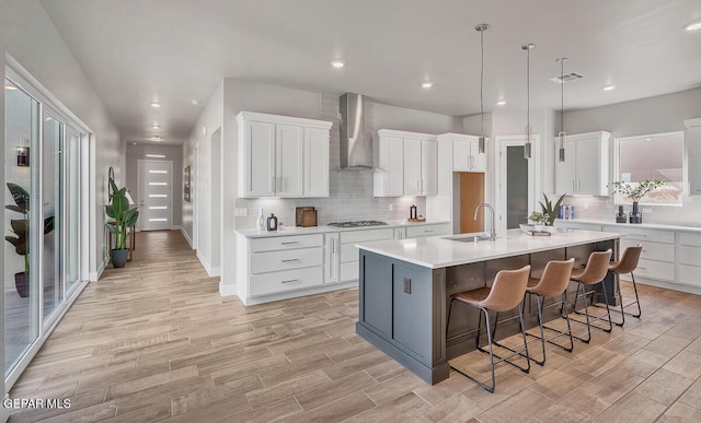 kitchen with wall chimney range hood, backsplash, an island with sink, white cabinets, and a breakfast bar