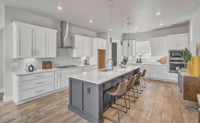 kitchen with backsplash, wall chimney exhaust hood, white cabinets, a kitchen island with sink, and appliances with stainless steel finishes