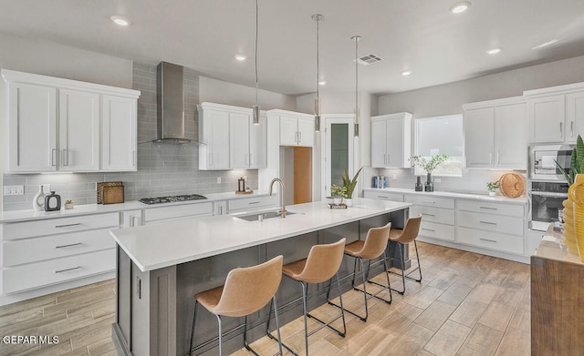 kitchen with wall chimney range hood, an island with sink, appliances with stainless steel finishes, sink, and tasteful backsplash