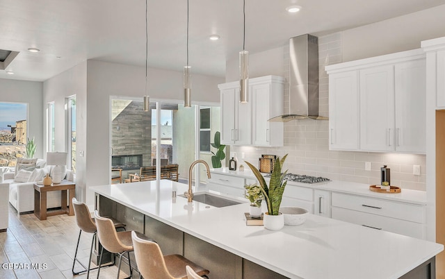 kitchen with a center island with sink, a fireplace, pendant lighting, wall chimney exhaust hood, and white cabinetry