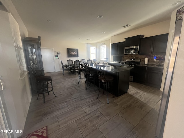 kitchen featuring an island with sink, stainless steel appliances, backsplash, dark tile flooring, and a breakfast bar area