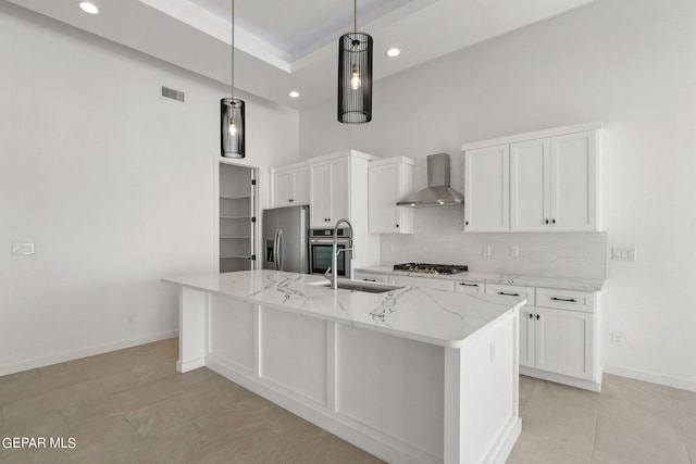 kitchen with an island with sink, decorative light fixtures, wall chimney range hood, white cabinetry, and appliances with stainless steel finishes