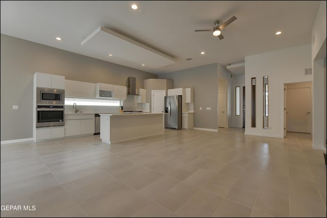 kitchen featuring ceiling fan, wall chimney range hood, a kitchen island, white cabinets, and appliances with stainless steel finishes