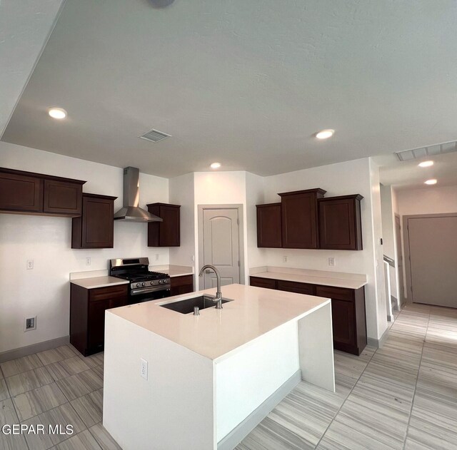 kitchen featuring dark brown cabinetry, sink, wall chimney exhaust hood, stainless steel gas range, and a kitchen island with sink