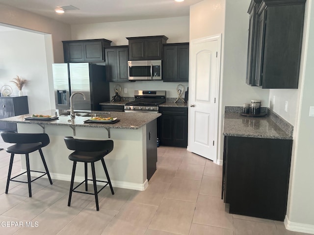 kitchen with stainless steel appliances, stone countertops, a kitchen island with sink, and dark cabinetry