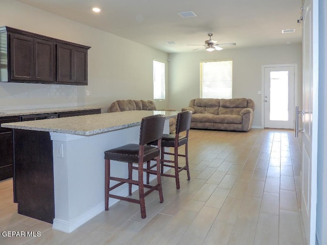 kitchen with ceiling fan, a center island, a kitchen breakfast bar, light stone counters, and dark brown cabinets