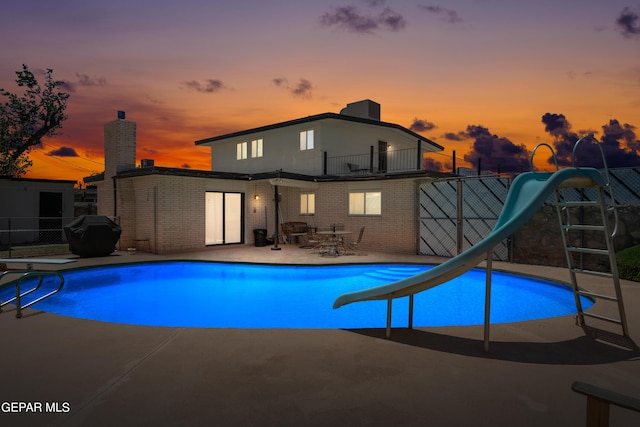 pool at dusk with a water slide, fence, a diving board, an outdoor pool, and a patio area