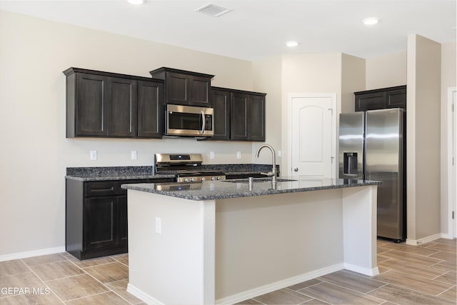 kitchen featuring sink, stainless steel appliances, dark stone counters, and a kitchen island with sink