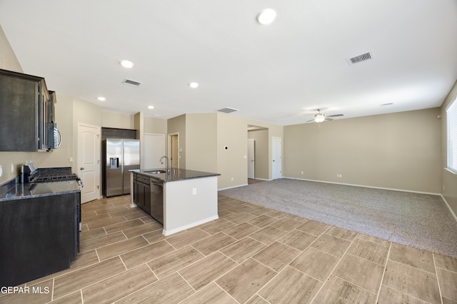 kitchen featuring stainless steel appliances, dark stone countertops, sink, ceiling fan, and a center island with sink