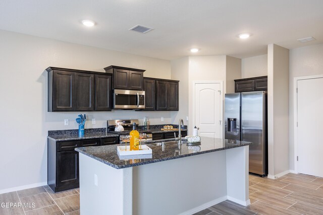 kitchen featuring a center island with sink, dark brown cabinets, sink, appliances with stainless steel finishes, and dark stone counters