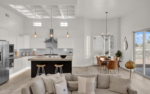 kitchen featuring wall chimney exhaust hood, coffered ceiling, an island with sink, beam ceiling, and decorative backsplash