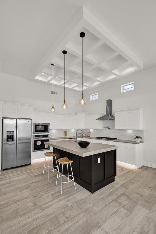 kitchen with white cabinetry, hanging light fixtures, a center island with sink, stainless steel appliances, and wall chimney range hood