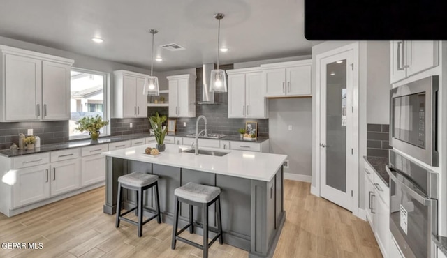 kitchen with wall chimney range hood, light hardwood / wood-style floors, appliances with stainless steel finishes, and a center island with sink