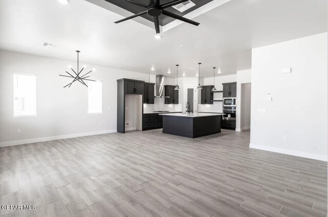 kitchen featuring wall chimney range hood, ceiling fan with notable chandelier, a kitchen island with sink, light hardwood / wood-style floors, and pendant lighting