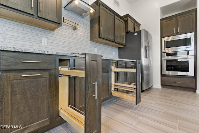 kitchen with tasteful backsplash, light stone counters, dark brown cabinetry, wall chimney exhaust hood, and stainless steel appliances