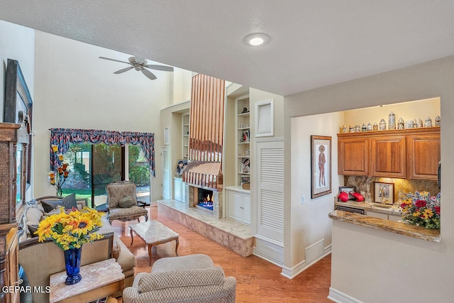 living room with a textured ceiling, light wood-type flooring, built in features, ceiling fan, and a fireplace