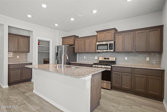 kitchen with dark brown cabinetry, sink, a center island with sink, stainless steel appliances, and light stone countertops