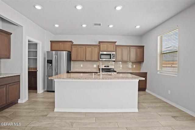 kitchen with light stone counters, stainless steel appliances, a center island with sink, and backsplash