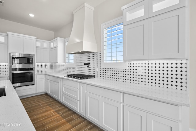 kitchen with custom exhaust hood, stainless steel appliances, decorative backsplash, wood tiled floor, and white cabinets