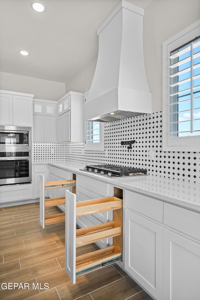 kitchen with tasteful backsplash, custom range hood, wood tiled floor, stainless steel appliances, and white cabinetry