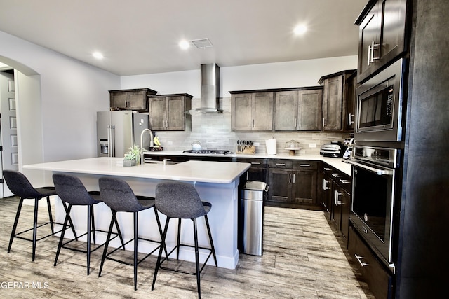 kitchen with a sink, appliances with stainless steel finishes, light wood-style flooring, wall chimney range hood, and a kitchen island with sink
