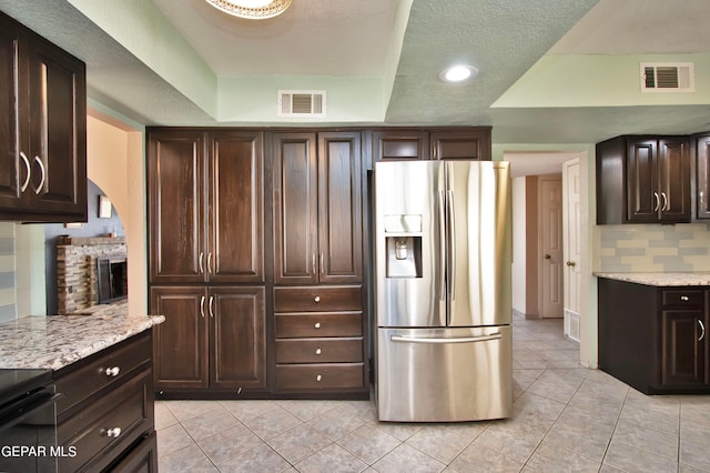 kitchen with light stone counters, stainless steel refrigerator with ice dispenser, a brick fireplace, a tray ceiling, and tasteful backsplash