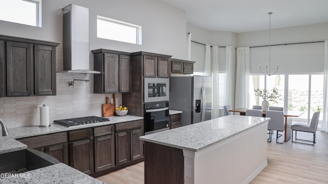 kitchen featuring appliances with stainless steel finishes, a notable chandelier, decorative light fixtures, wall chimney exhaust hood, and light stone countertops