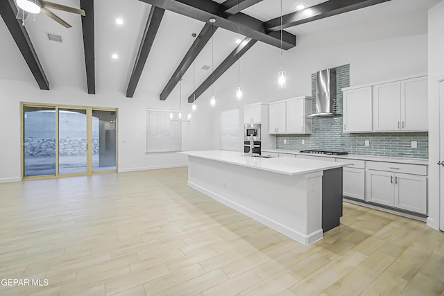 kitchen with beam ceiling, stainless steel appliances, hanging light fixtures, wall chimney range hood, and light hardwood / wood-style flooring