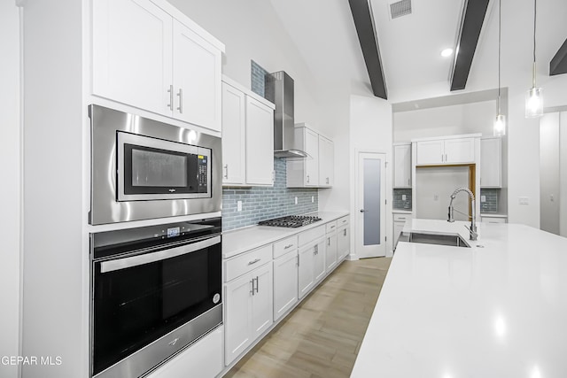 kitchen with appliances with stainless steel finishes, white cabinets, wall chimney exhaust hood, beamed ceiling, and hanging light fixtures