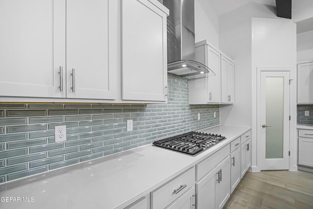 kitchen with wall chimney exhaust hood, backsplash, light hardwood / wood-style floors, stainless steel gas stovetop, and white cabinets