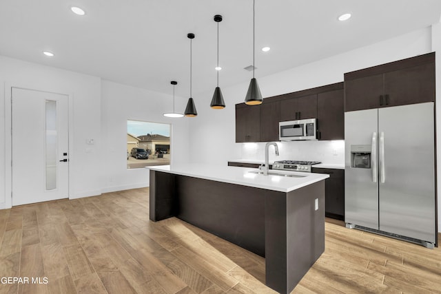 kitchen with light wood-type flooring, stainless steel appliances, an island with sink, decorative backsplash, and decorative light fixtures