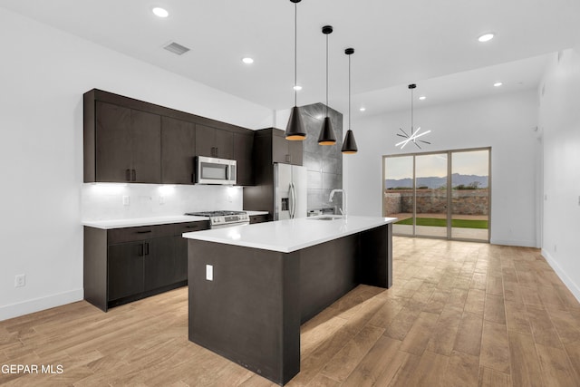 kitchen featuring tasteful backsplash, a center island with sink, stainless steel appliances, light hardwood / wood-style flooring, and decorative light fixtures