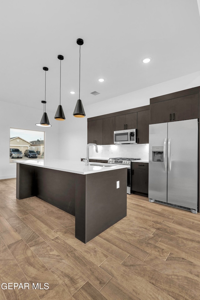 kitchen featuring light hardwood / wood-style flooring, stainless steel appliances, a kitchen island with sink, hanging light fixtures, and tasteful backsplash