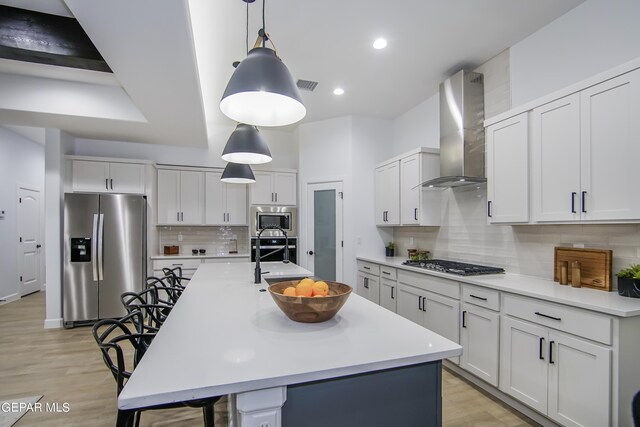 kitchen with white cabinets, an island with sink, decorative light fixtures, wall chimney exhaust hood, and appliances with stainless steel finishes