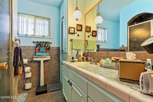 bathroom featuring a stall shower, a wainscoted wall, stone tile flooring, vanity, and tile walls