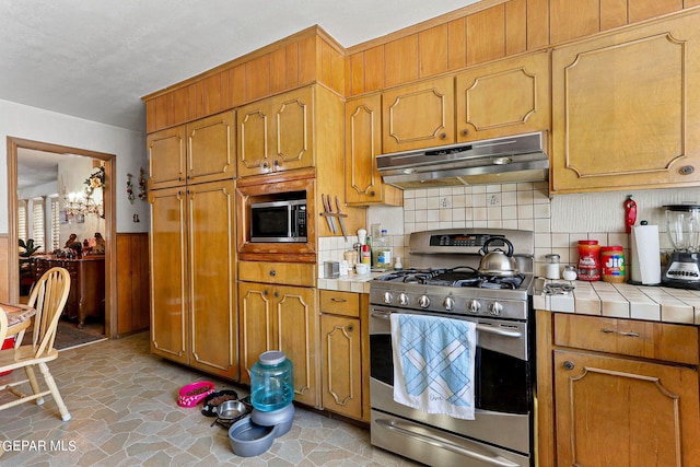 kitchen with brown cabinetry, tile countertops, stone finish floor, stainless steel appliances, and under cabinet range hood