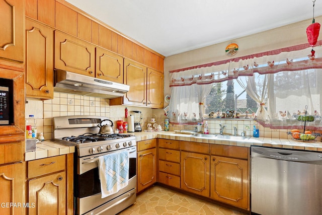 kitchen with tasteful backsplash, tile countertops, stainless steel appliances, under cabinet range hood, and a sink