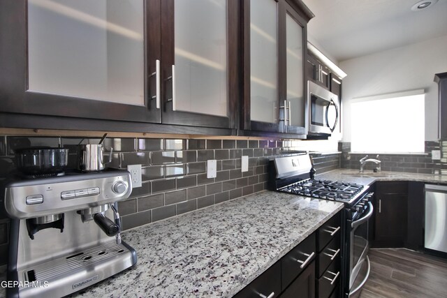 kitchen with light stone counters, dark brown cabinetry, appliances with stainless steel finishes, backsplash, and glass insert cabinets