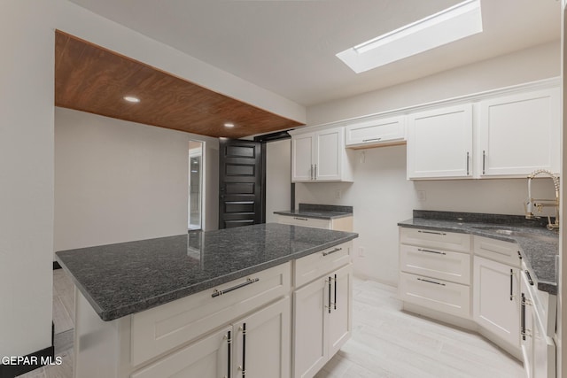 kitchen featuring light wood-style flooring, a sink, white cabinetry, dark stone counters, and a skylight