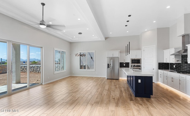 kitchen with appliances with stainless steel finishes, light hardwood / wood-style floors, decorative backsplash, an island with sink, and wall chimney range hood