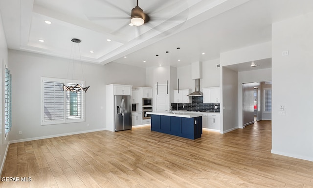 kitchen with wall chimney range hood, pendant lighting, a tray ceiling, an island with sink, and stainless steel appliances