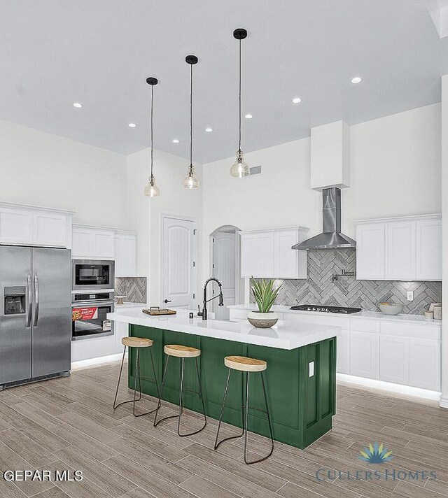 kitchen featuring tasteful backsplash, a large island with sink, stainless steel appliances, wall chimney range hood, and white cabinets