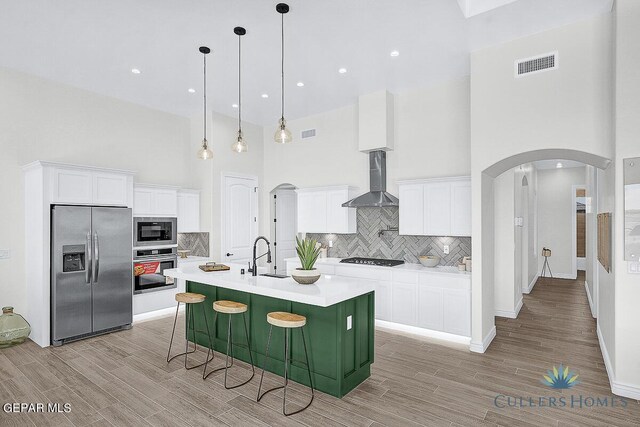 kitchen with wall chimney range hood, backsplash, white cabinetry, appliances with stainless steel finishes, and a center island with sink